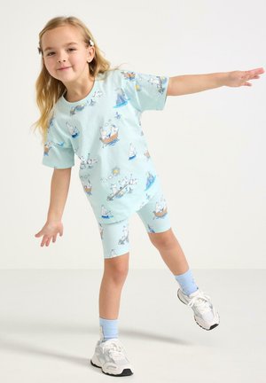 Młoda dziewczyna balansująca na jednej nodze, ubrana w jasnoniebieską koszulkę z nadrukiem Muminka, pasujące kolarki, niebieskie skarpetki i białe trampki.