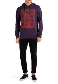 Sudadera morada con un estampado gráfico rojo que dice “JACK & JONES,” combinada con pantalones negros y zapatillas negras. Tejido suave con un ajuste relajado.