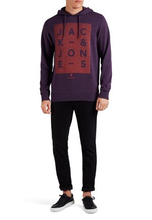Sudadera morada con un estampado gráfico rojo que dice “JACK & JONES,” combinada con pantalones negros y zapatillas negras. Tejido suave con un ajuste relajado.