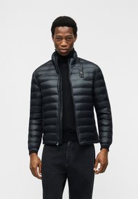 Άνδρας που φοράει μαύρο μπουφάν τύπου puffer πάνω από μαύρο ζιβάγκο και μαύρο παντελόνι, στέκεται μπροστά σε απλό φωτεινό φόντο.