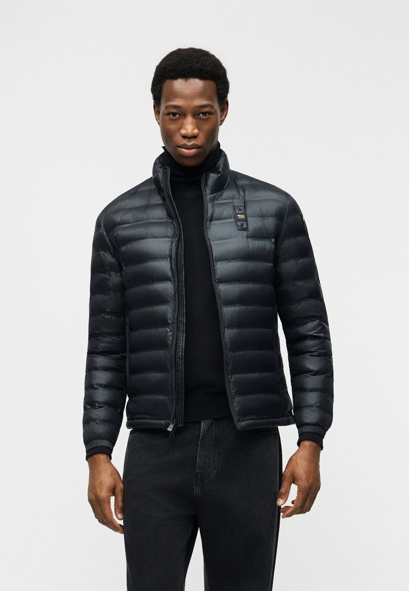 Άνδρας που φοράει μαύρο μπουφάν τύπου puffer πάνω από μαύρο ζιβάγκο και μαύρο παντελόνι, στέκεται μπροστά σε απλό φωτεινό φόντο.