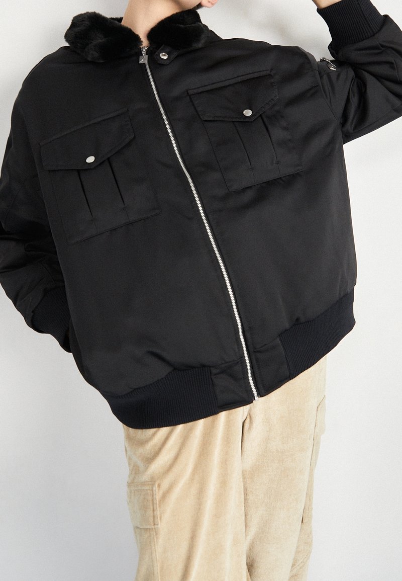 Veste noire zippée avec deux grandes poches à rabat et un col doublé de fourrure portée avec un pantalon beige.