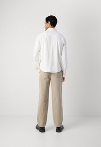 Witte langesleeve shirt met een gebogen onderrand, gecombineerd met losse beige broek en zwarte schoenen. Eenvoudig ontwerp zonder zichtbare patronen of accenten.