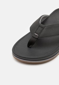 Musta flip-flop, jossa on teksturoitu synteettinen hihna, kumipohja ja minimaalinen harmaa yksityiskohta. Sisältää varvaslenkin turvallisen istuvuuden ja mukavuuden takaamiseksi.