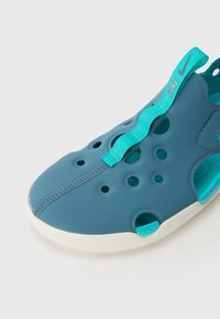 Teal slip-on kengät ilmanpitoa varten, sileä kumimateriaali, valkoinen pohja ja teal-väriinen hihna, jossa on logo sivulla.