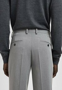 Szare spodnie o dopasowanym kroju, z podwójnymi zakładkami i tylnymi kieszeniami zapinanymi na czarne guziki. Materiał wydaje się gładki, o drobnej fakturze.
