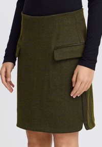 Jupe à motif chevron vert avec deux poches latérales, fabriquée à partir d'un tissu texturé. Ourlet ajusté, portée avec un haut noir.