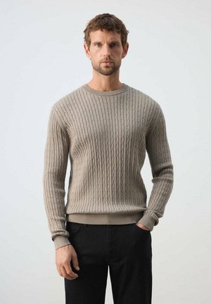 Man standing with neutral expression wearing a beige cable-knit sweater and black pants against a plain background.