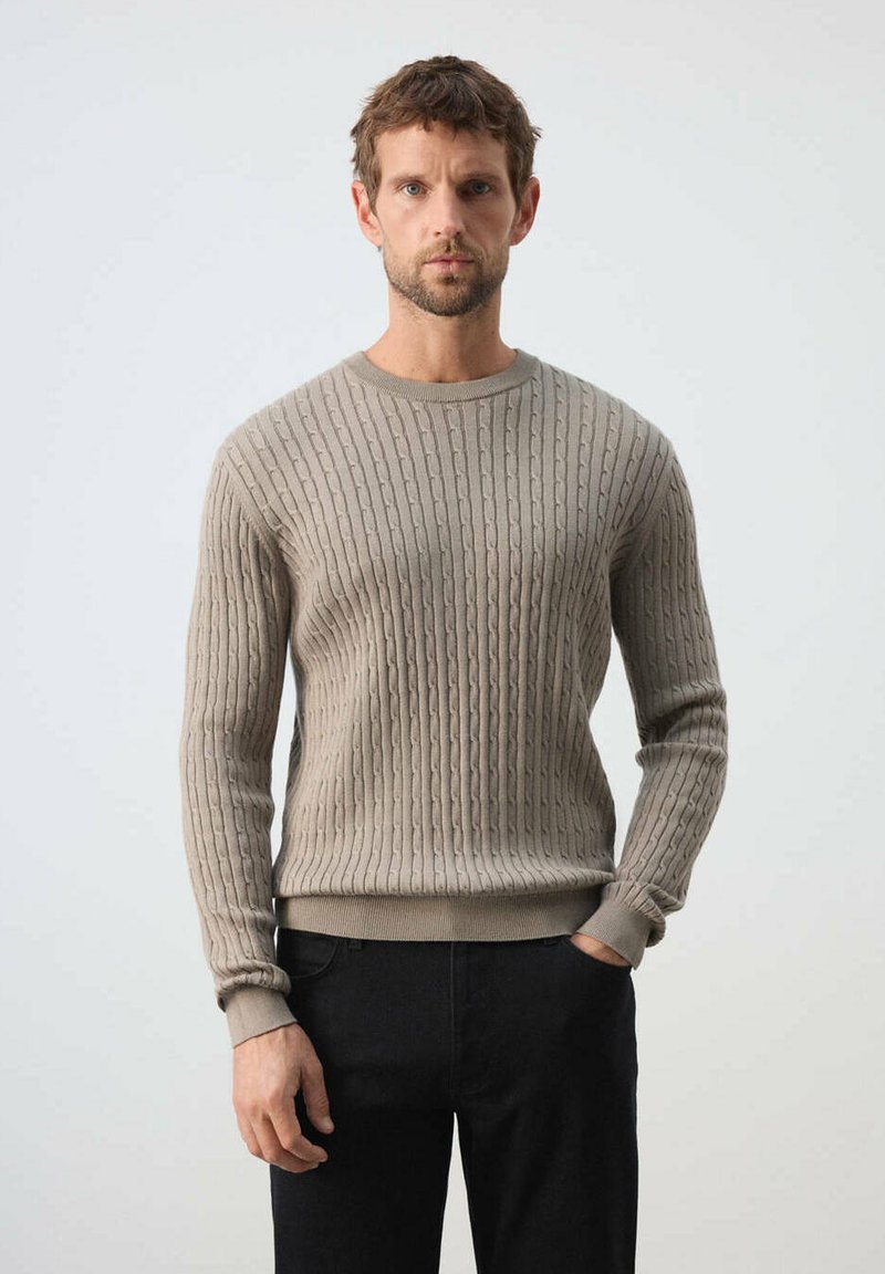 Homme debout avec une expression neutre, portant un pull beige à torsades et un pantalon noir sur un fond uni.