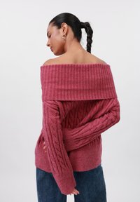 Off-the-shoulder, ribbed pink sweater with a cable knit design and thick cuffs, paired with dark blue denim jeans.