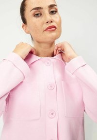 Ljusrosa kofta gjord av texturerat tyg, med en krage, två framfickor och stora knappar. Ärmarna är något överdimensionerade.