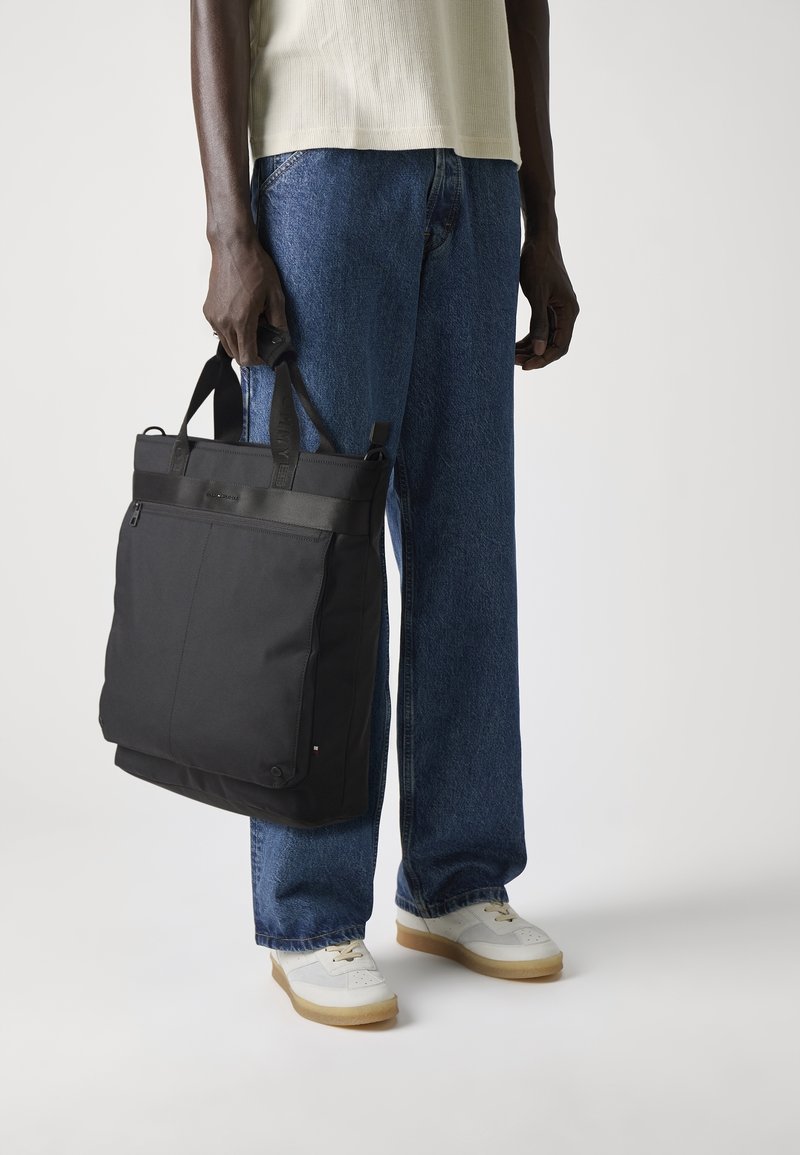 Personne portant un jean bleu et des baskets blanches tenant un grand sac cabas noir par ses anses sur fond blanc.