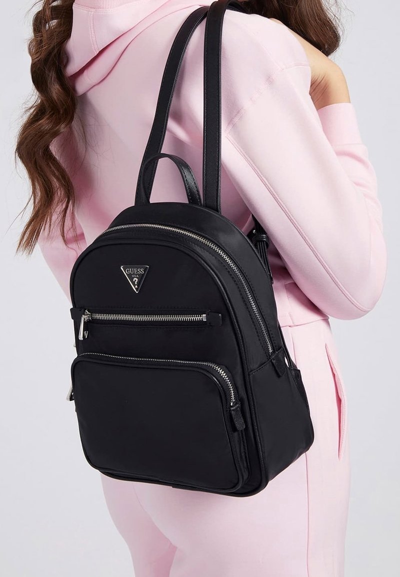 Femme portant un sweat à capuche rose clair et un pantalon, portant un sac à dos noir Guess avec des fermetures éclair argentées sur l'épaule.