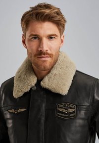 Chaqueta de cuero negra con un cuello de borreguito beige. Presenta un parche de emblema en el pecho y detalles alados en los hombros.