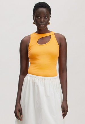 Woman with short hair wearing a sleeveless orange top with an asymmetrical cutout and a white skirt, standing against a light background.