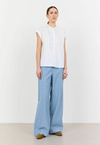 Camicia bianca a maniche corte con dettagli arricciati sul davanti, abbinata a pantaloni a gamba larga blu chiaro e scarpe piatte marroni su uno sfondo neutro.