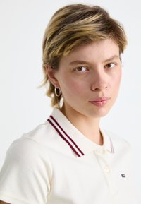 Polo shirt couleur crème avec un tissu texturé, mettant en avant un col à rayures bordeaux bicolores et un petit logo sur la poitrine.