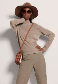 Beige gestrickter Pullover mit gerippter Textur, brauner Hut mit breiter Krempe, übergroße Sonnenbrille und eine braune Umhängetasche. Glatte, strukturierte Hose.