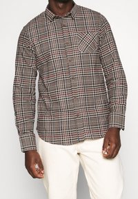 Hombre vestido con una camisa de cuadros en tonos marrón, negro y rojo, con un bolsillo en el pecho, y pantalones de color crema, de pie frente a un fondo liso.