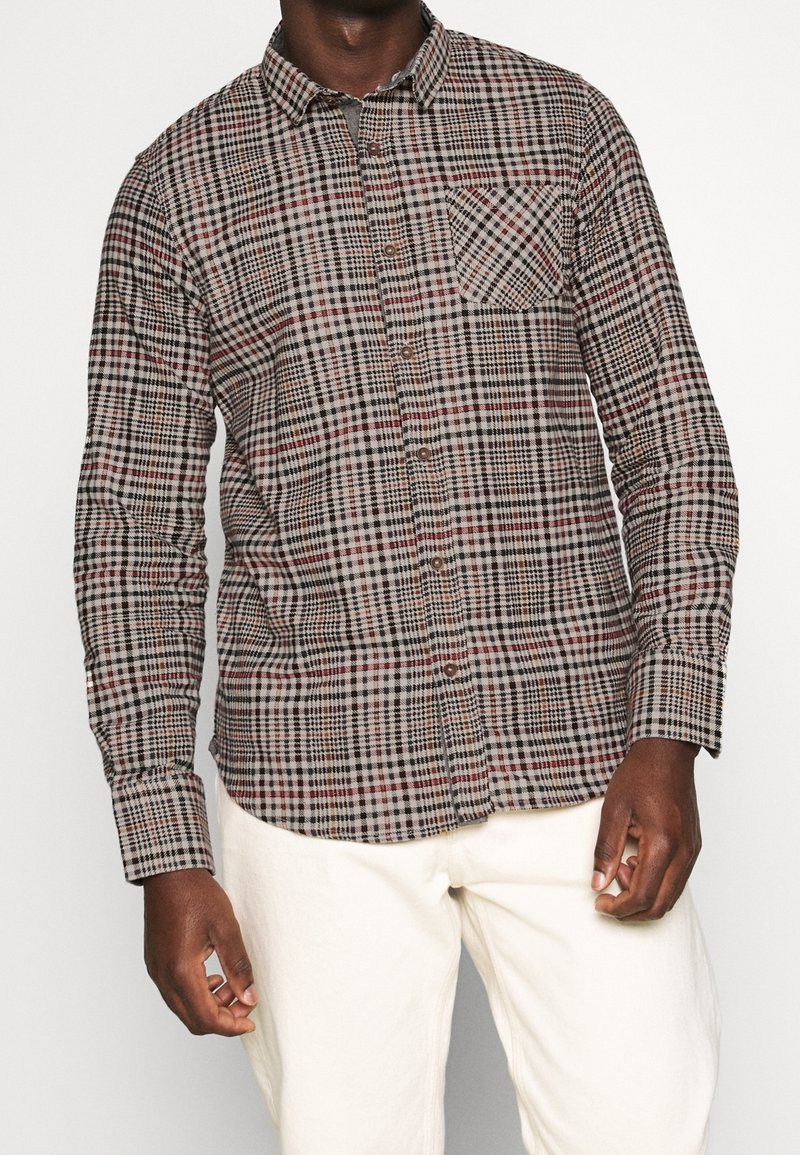 Hombre vestido con una camisa de cuadros en tonos marrón, negro y rojo, con un bolsillo en el pecho, y pantalones de color crema, de pie frente a un fondo liso.