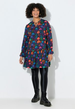 Kvinde med krøllet hår smilende, iført en farverig blomsterkjole, sorte leggings og sorte snørestøvler, stående mod en hvid baggrund.