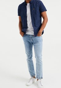 Krátký rukáv námořnicky modré vzorované košile přes bílé tričko, doplněné světle modrými potrhanými džínami a bílými teniskami.