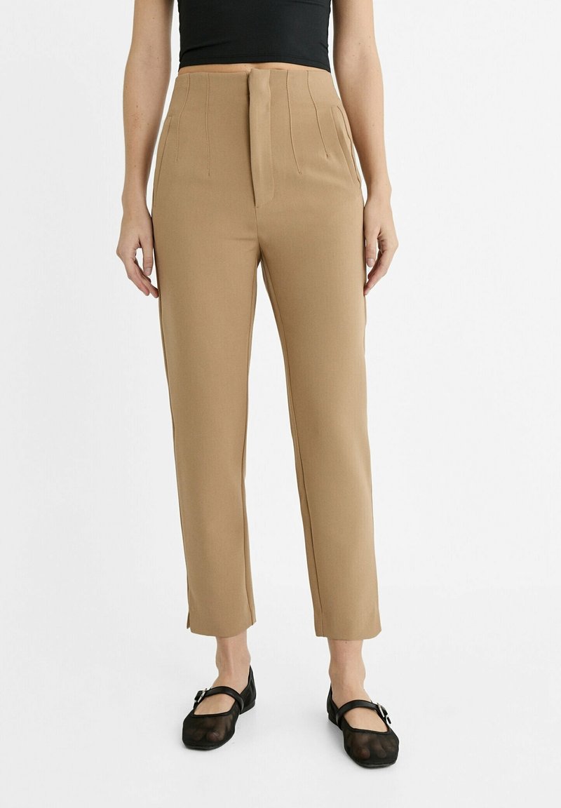 Femme portant un pantalon beige ajusté taille haute, des chaussures Mary Jane noires transparentes et un haut noir sans manches.