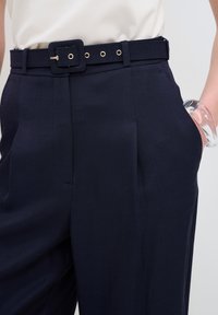 Pantalons taille haute bleu marine avec des plis, dotés d'une large ceinture avec une boucle carrée et des œillets en ton doré ; texture de tissu lisse.
