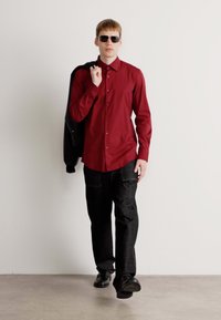 Uomo che indossa occhiali da sole, camicia rossa a bottone, pantaloni neri e giacca nera sulle spalle, che cammina in avanti su uno sfondo neutro.