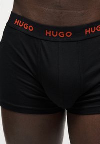 Μαύρα ανδρικά boxer briefs με μαύρη ελαστική μέση που φέρει το όνομα της μάρκας "HUGO" με κόκκινα γράμματα.