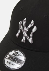 Schwarze Kappe aus Stoff mit grau gesticktem Logo und verstellbarem Riemen. Zeigt ein metallisches Etikett mit Marken- und Modellinformationen.