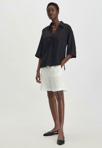 Camicia nera in lino con colletto e maniche corte, abbinata a pantaloni corti bianchi in lino. Il modello indossa delle ballerine nere; taglio rilassato e colori neutri.