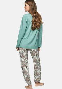 Vrouw met lang haar die een mintgroene langslijfs top en witte met paisleypatroon bedrukte broek draagt, staand op blote voeten tegen een witte achtergrond.