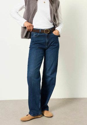 Person trägt blaue Jeans mit braunem Gürtel, weißes Langarmhemd, graue Weste und beige Slipper und steht drinnen.