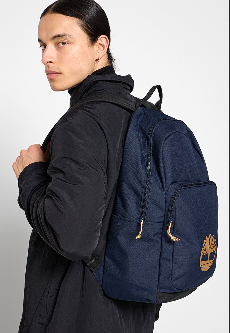 Sac à dos bleu marine en tissu texturé, avec des bretelles réglables, des détails en matériel orange et un grand logo doré d'arbre sur le côté.