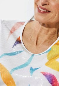 Weißes Pailletten-Oberteil mit rundem Ausschnitt, das farbenfrohe abstrakte Muster in Blau, Orange, Gelb und Lila zeigt. Strukturelles Detail sichtbar.