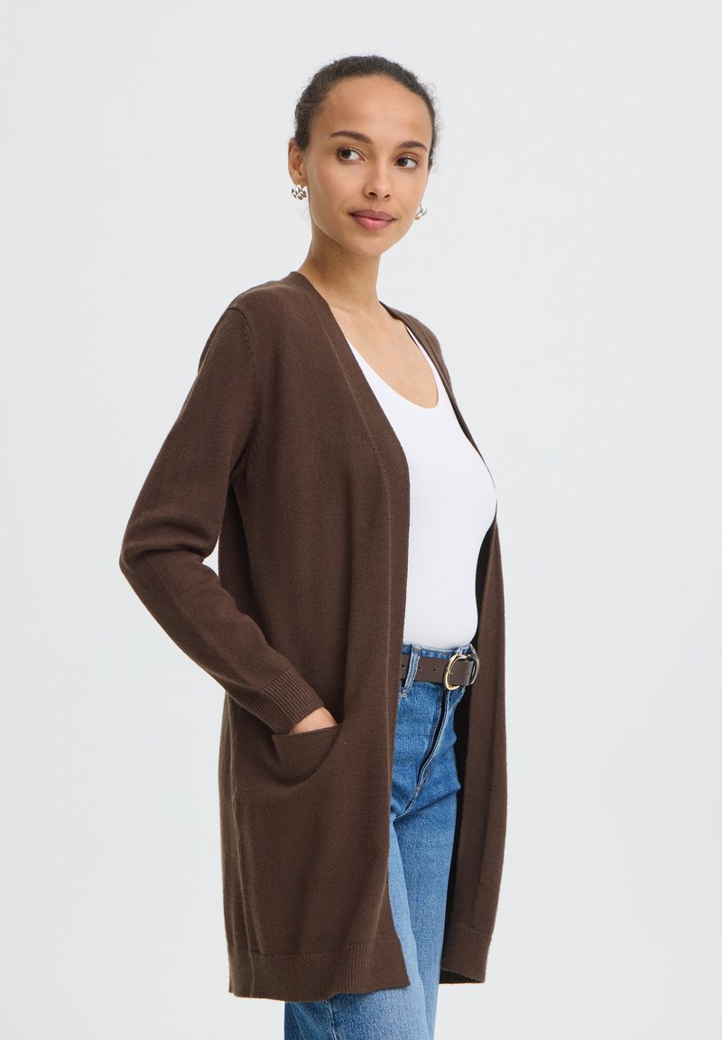 Femme aux cheveux attachés, portant un cardigan marron ouvert sur un haut blanc et un jean bleu avec ceinture, debout devant un fond clair uni.