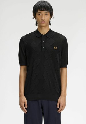 Giovane uomo che indossa una polo nera a maniche corte con motivo diamantato strutturato e piccolo logo d’alloro dorato sul petto.