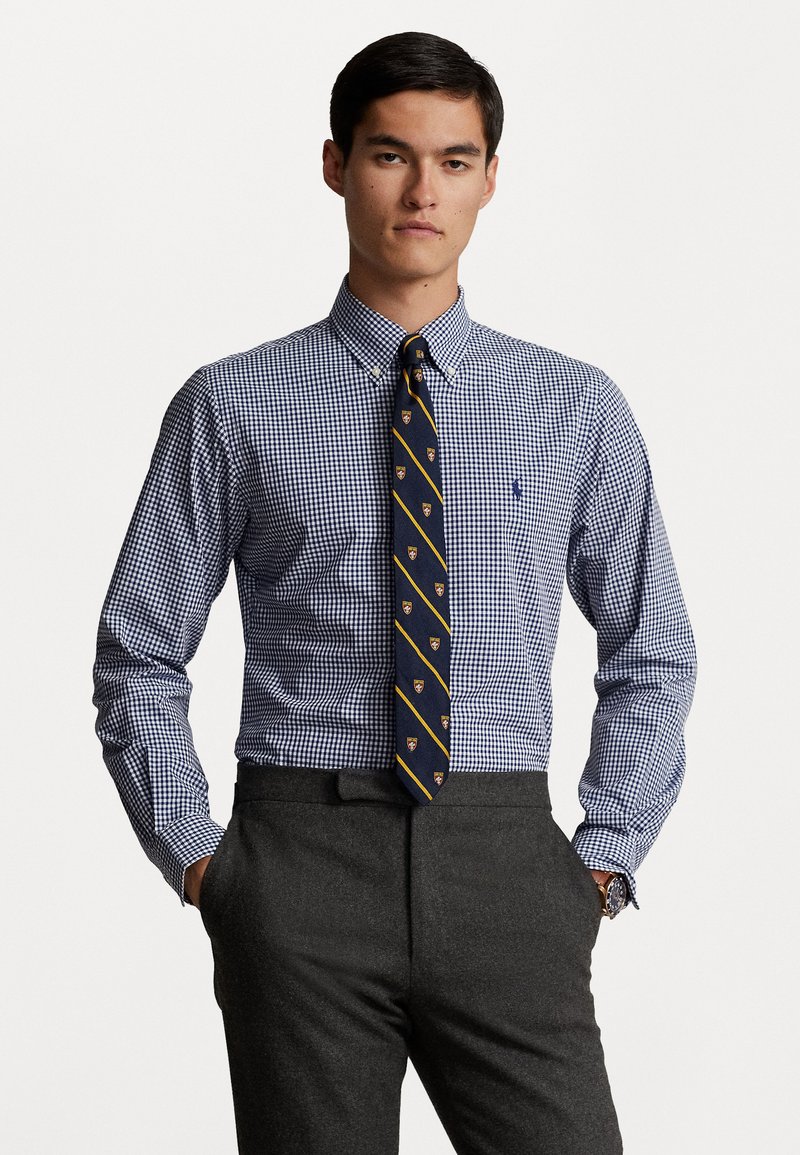 Navy en witte gingham dress shirt met button-down kraag, gecombineerd met een navy stropdas met gele strepen en logo's, gedragen met grijze broeken.