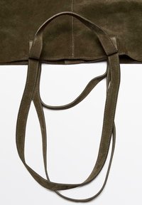Suède handtas in olijfgroen met een gladde textuur, voorzien van lange banden en zichtbare stikdetails. Het ontwerp benadrukt minimalistische esthetiek.