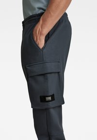 Pantalones de chándal gris oscuro con un bolsillo lateral, fabricados con un tejido texturizado y una cintura elástica. Se puede ver un parche de logo negro en el dobladillo.