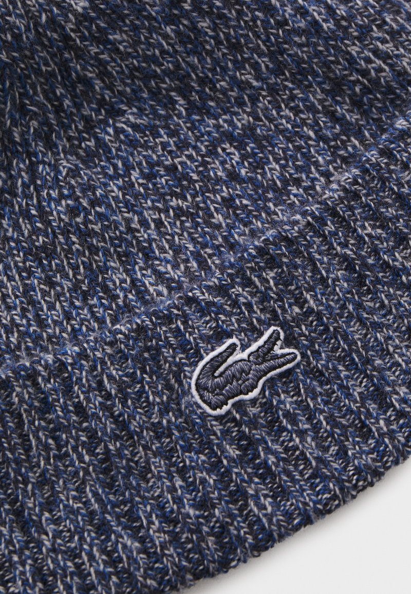Bonnet en tricot dans des nuances de bleu avec un motif texturé. Présente un logo brodé blanc sur le bord. Le matériau semble doux et chaud.