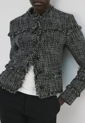 Personne portant une veste en tweed noir et blanc avec des bords frangés et des boutons noirs, par-dessus un haut blanc et un pantalon noir.