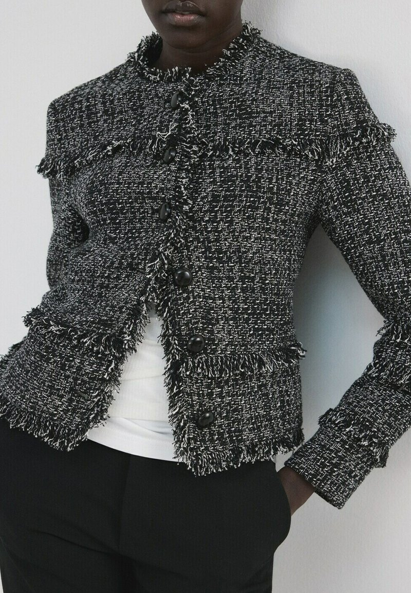 Personne portant une veste en tweed noir et blanc avec des bords frangés et des boutons noirs, par-dessus un haut blanc et un pantalon noir.