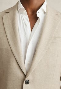 Blazer texturat bej cu rever ascuțit, un singur nasture negru, purtat peste o cămașă albă din in cu nasturi. Design simplu și modern.