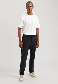 Witte kortebloes met een zak, gecombineerd met zwarte broek met trekkoord. Het model draagt beige en witte sneakers. Gladde stofstructuren.