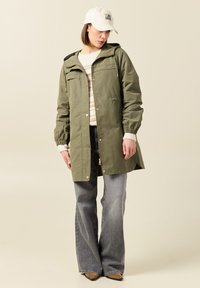 Parka vert olive avec capuche, fermeture éclair et boutons, deux poches poitrine, poignets élastiques. Portée sur un pull rayé et un jean évasé.
