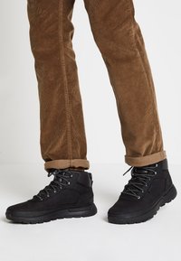 Bottes de randonnée noires avec un dessus texturé, un design à lacets et des semelles en caoutchouc, associées à un pantalon en velours côtelé marron retroussé.