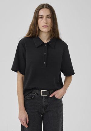 Jeune femme aux longs cheveux bruns portant une chemise noire à manches courtes avec col, rentrée dans un jean noir avec une ceinture noire, une main dans la poche.