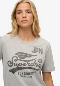 T-shirt gris à manches courtes avec un grand logo graphique noir présentant un design d'aile, "Superdry," et des éléments de texte dans un style vintage.